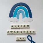 Paper cutout rainbow and tiles form 'Autism Awareness Day' with a blue ribbon on a light background.