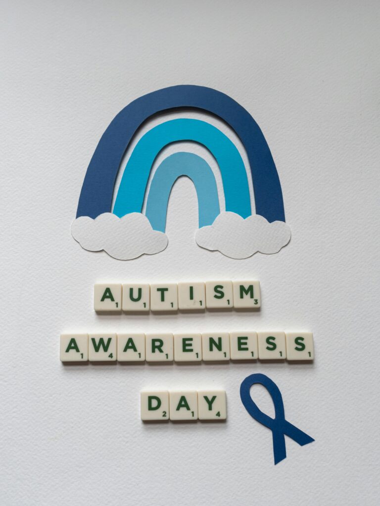 Paper cutout rainbow and tiles form 'Autism Awareness Day' with a blue ribbon on a light background.